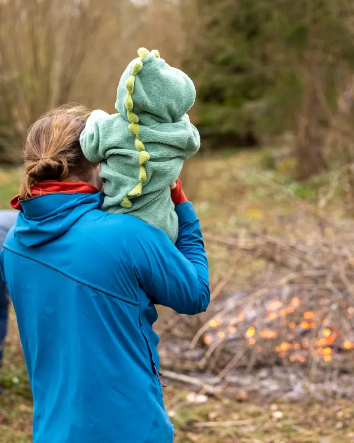A parent carrying a baby in a green dinosaur hood by a bonfire, seen from behind