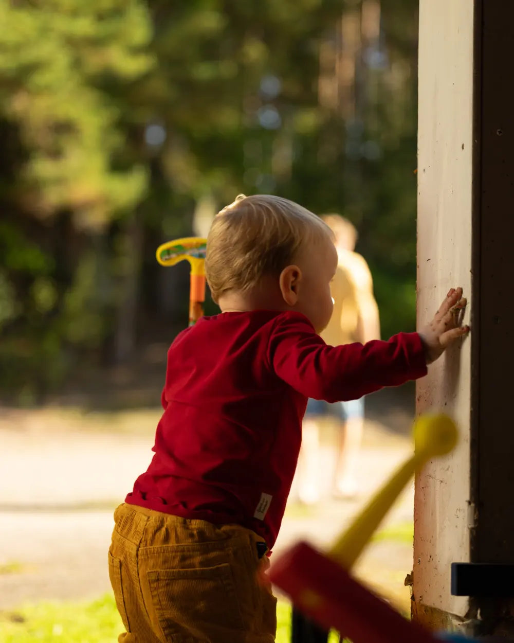 A toddler in a red shirt leaning on a doorpost in the evening sun, seen from behind